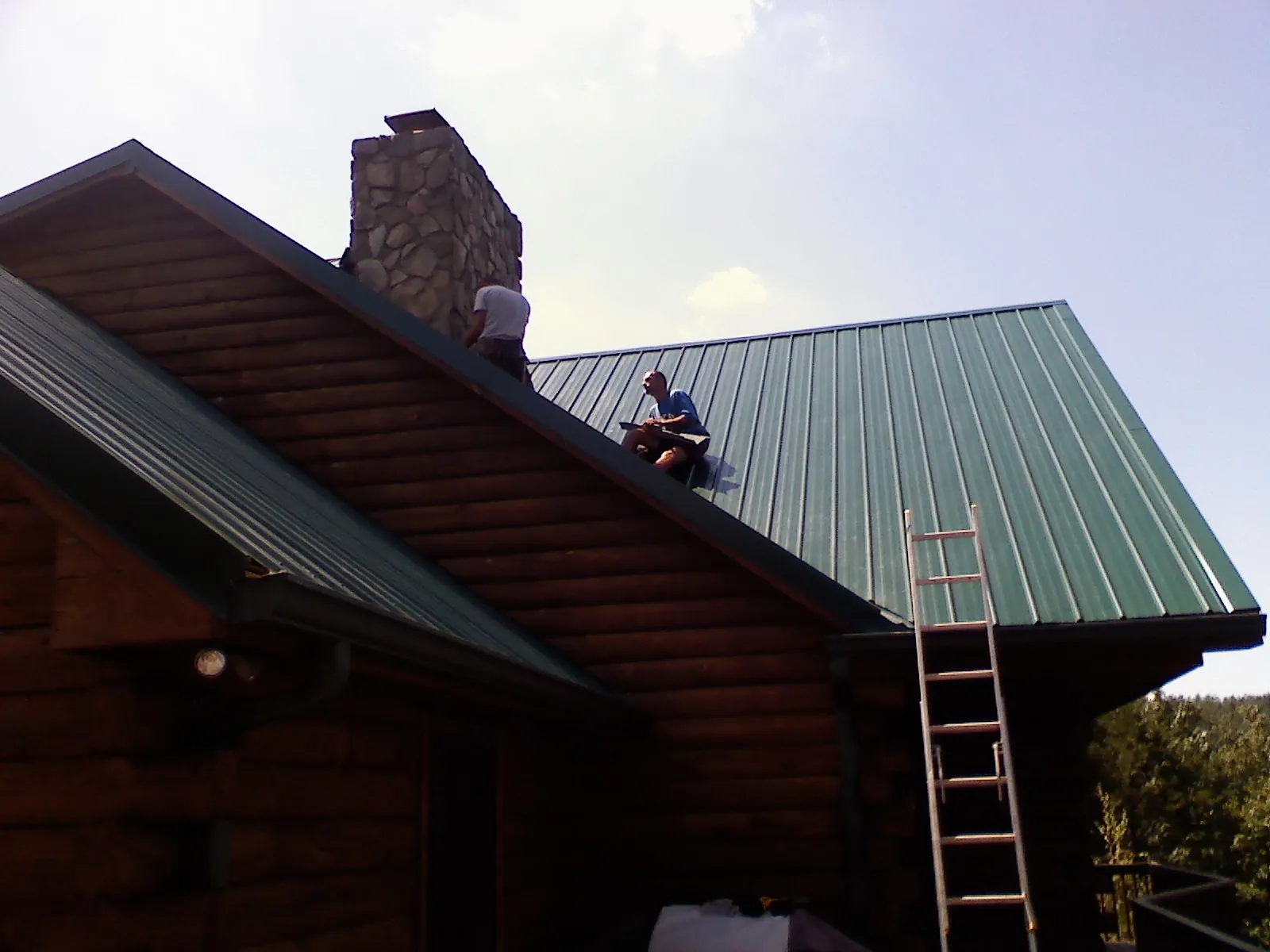 Roofers installing green metal panels beside a stone chimney on a log home in Erie
